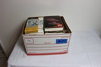 Cardboard box filled with miscellaneous used books, visible titles include 'The Hearing' and 'Signs of the Times'.