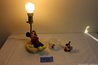 Mickey Mouse nursery lamp nightlight turned on showing illumination with seated Mickey base, milk glass mug and bisque figurine placed beside it