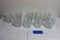 Nine vintage clear glass mugs with a dimpled thumbprint pattern arranged in two rows on a white cloth background.