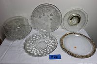 Wide shot of all glassware items on white cloth showing crystal pedestal cake stand with dome, three large plates including Heisey and Mikasa platters, and unbranded platter with gold rim.