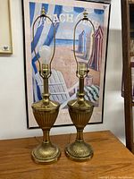 Two brass table lamps on a wooden surface in front of a framed print. Lamps feature ribbed fluted bases and bodies, brass harp arms, and light bulb sockets.