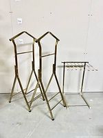 Full view of two brass-tone metal clothing racks for suits or dresses and one smaller rack for pants with horizontal rods, displayed against a plain white wall.