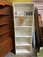 Front view of white IKEA Billy bookcase filled with glassware to show shelving structure and condition. Nearby furniture pieces visible.