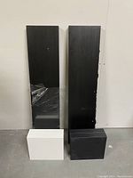 Two large black shelves standing vertically with one partially wrapped in plastic with some visible scratches. Two white shelves and one smaller black shelf placed in front on the floor.