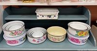 Photo showing nine nesting bowls and containers with various floral patterns placed on a shelf.