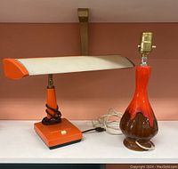 Photo of both lamps side by side on shelf with orange desk lamp on left and mid century modern ceramic drip glaze table lamp on right