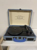 Front view of the portable record player open, showing the turntable, tonearm, control knobs, and interior lid branding.