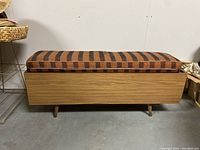 Side view of rectangular wooden storage bench with striped cushion on top. Bench has mid-century style tapered legs and wood veneer finish.