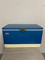 Front view of a retro blue Coleman cooler with metal latch, white bottom, and logo visible.