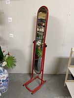 Front view of a red floor-length oval mirror with metal frame and black adjustment knobs on the sides, standing on a rounded rectangular base with black feet.
