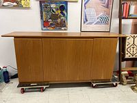 Full front view of vintage teak wood credenza on dollies indoors with artwork in background.