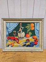 Full view of framed vintage cross stitch depicting lobster, wine bottle, glass, and fruits