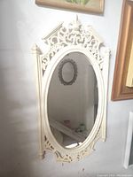 Full view of the vintage wall mirror showing the oval glass and ornate white wooden frame with scroll and floral designs.
