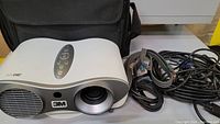 3M S10 projector shown with lens, vent, and control buttons on top sitting beside a carrying bag.