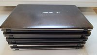 Five stacked Asus laptops closed showing black lids with Asus logo, indicating quantity and model range