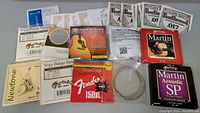 Photo showing assortment of banjo and guitar string packages from multiple brands such as Martin, Vega, Fender, Long & McQuade, and Newtone spread out on a surface.