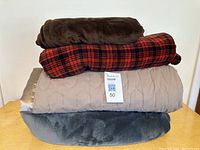 Stacked view of four blankets showing brown, red plaid, beige with satin edging, and grey plush blankets.