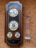 Full front view of the Bulova weather station on dark wood panel and the glass Galileo thermometer standing side by side on the carpet.