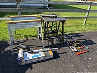 Overall lot showing two metal saw horses with wooden tops on left, two black Jobmate foldable work benches on right, various clamps laid out on ground, and paint sticks with nail puller in front.
