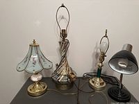 Four table lamps displayed on a table against a white wall, including floral, vintage turquoise, brass-green, and black desk lamps.