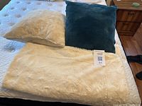Cream-colored plush throw blanket spread out with matching cream cushion and one green velvet pillow on top showing size and texture.