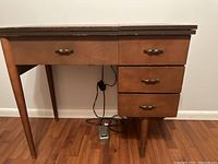 Photo of closed wooden sewing cabinet with three drawers on the right side and tapered legs. Power cord and foot pedal visible under the cabinet.