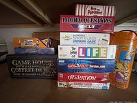 Photo showing stacked board games including Loaded Questions Adult, Shot Glass Basketball Drinking Game, The Game of Life, Monopoly Version.com, Operation, Candy Land, and a jungle-themed game.