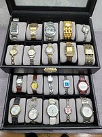 Top-down view of 18 wristwatches arranged in a black watch box with grey cushioned compartments.