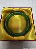 Green jade bangle bracelet resting inside a red and yellow satin-lined presentation box, showing the full shape and polish of the bracelet.