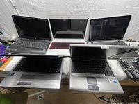 Five laptops placed on a table, shown from the top angle; includes black, gray, white/red colors, power cords visible on side