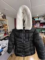 Full front view of black puffer winter coat with hood on a dress form in a room with shelves.