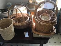 Photo showing collection of nine wicker baskets of varying sizes, shapes and handles arranged on a table.