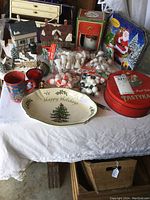 Wide view of mixed Christmas lot featuring ceramic village houses, tins, a holiday serving plate, and craft items like mini marshmallows and small snowmen.