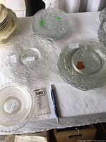 Top view of assorted clear glass plates showing different patterns including grape clusters and floral designs, arranged on a white tablecloth with a pen for scale.