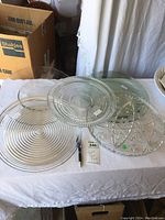 Four large circular glass trays displayed on a white cloth-covered surface, includes one transparent green tray and three clear glass trays with various designs.