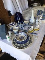 Collection of blue and white porcelain items including teapots, vases, figurines, plates, salt and pepper shakers, and decorative pieces arranged on a white cloth.