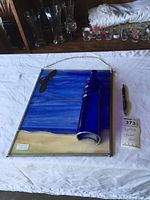 Full view of the rectangular stained glass panel featuring a blue 3D lighthouse on a blue and beige background, laid on white cloth with a pen for scale.