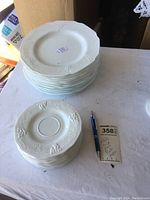Stack of 10 white milk glass dinner plates with embossed grape clusters on the rim seen from above, alongside a smaller stack of 10 matching saucers with the same grape motif.