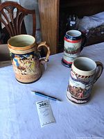 Overview of three painted beer steins shown on a white cloth, displaying detailed artwork and branding.
