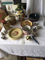 View of various pottery and ceramic items on white tablecloth, including multi-spout jug, plates, lidded jar, silver-tone bird figurines, and small floral figurine.