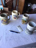 Image showing four ceramic steins on a white cloth. Steins feature traditional German and European designs, including colorful scenes, emblems, and floral patterns.