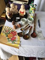 Full lot overview showing sumo figurines, male and female figurines, sake cups, wooden eggs, painted wooden spoons, and floral folding fan.