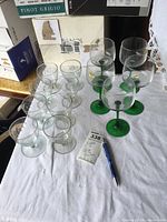 Full view of the set of glasses on a white tablecloth, with 5 larger wine glasses with green stems and 7 smaller clear stemmed glasses grouped together.