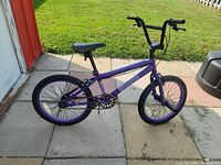 Side view of purple BMX bike with black tires and pegs, handlebars and seat visible against outdoor patio background.