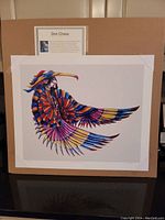 Full view of the unframed lithograph print showing the vibrant depiction of a Native American figure in colorful bird-like attire with a solid white background. The photo also shows a separate artist biography card above the print.