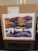 Photo showing the full unframed print laid on a brown backing board with a certificate of authenticity above the print. The artwork depicts hills and trees reflected on a lake in warm purples, oranges, and blues.