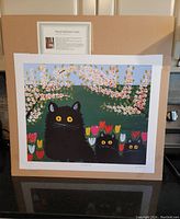 Full view of the unframed lithograph showing three black cats among flowers and blossoms, with a label describing Maud Lewis.