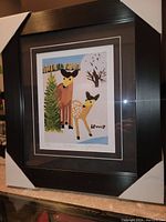 Full view of the professionally framed 'Two Deer' art print by Maud Lewis showing the black frame with white and black double mat surrounding the print of two deer in a folk art style winter scene, artist signature visible.
