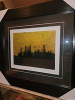 Framed fine art print of trees silhouetted against a textured yellow sky, titled 'Sunset Canoe Lake' by Tom Thomson. Black wood frame with white matting visible.
