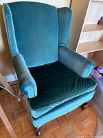 Green vintage velvet wingback armchair with rolled arms and wooden legs, showing one chair from angled side view.